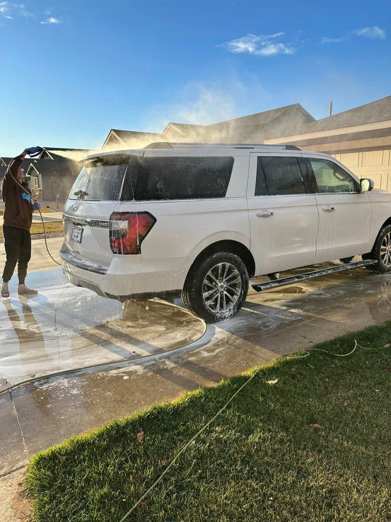 Technician pressure washing a white SUV in a residential driveway